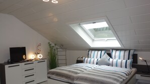 Un dormitorio con tragaluces, una cama con ropa de cama a rayas, un televisor en un mueble y una planta.