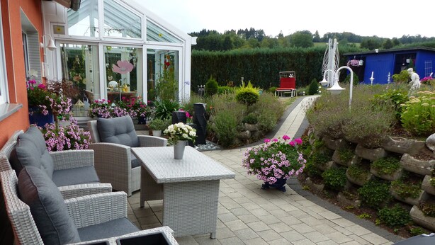 Un patio exterior con mobiliario de mimbre, plantas en macetas y una estructura de vidrio.