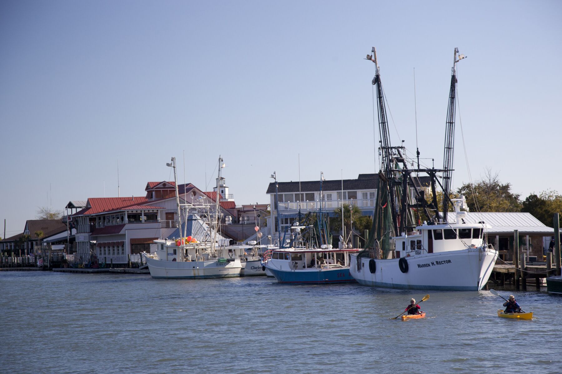 A marina with several boats docked, a building with a red roof, and kayakers on the water.