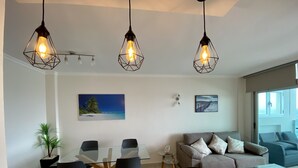 Ein modernes Wohnzimmer mit Essbereich, ausgestattet mit einem Glastisch, einer Couch und einem Gemälde mit einer Strandansicht.