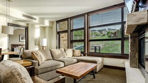 A modern living room with a sectional sofa, a wooden coffee table, and a fireplace. Large windows provide views of the outdoors. - Base Village (Snowmass Village)