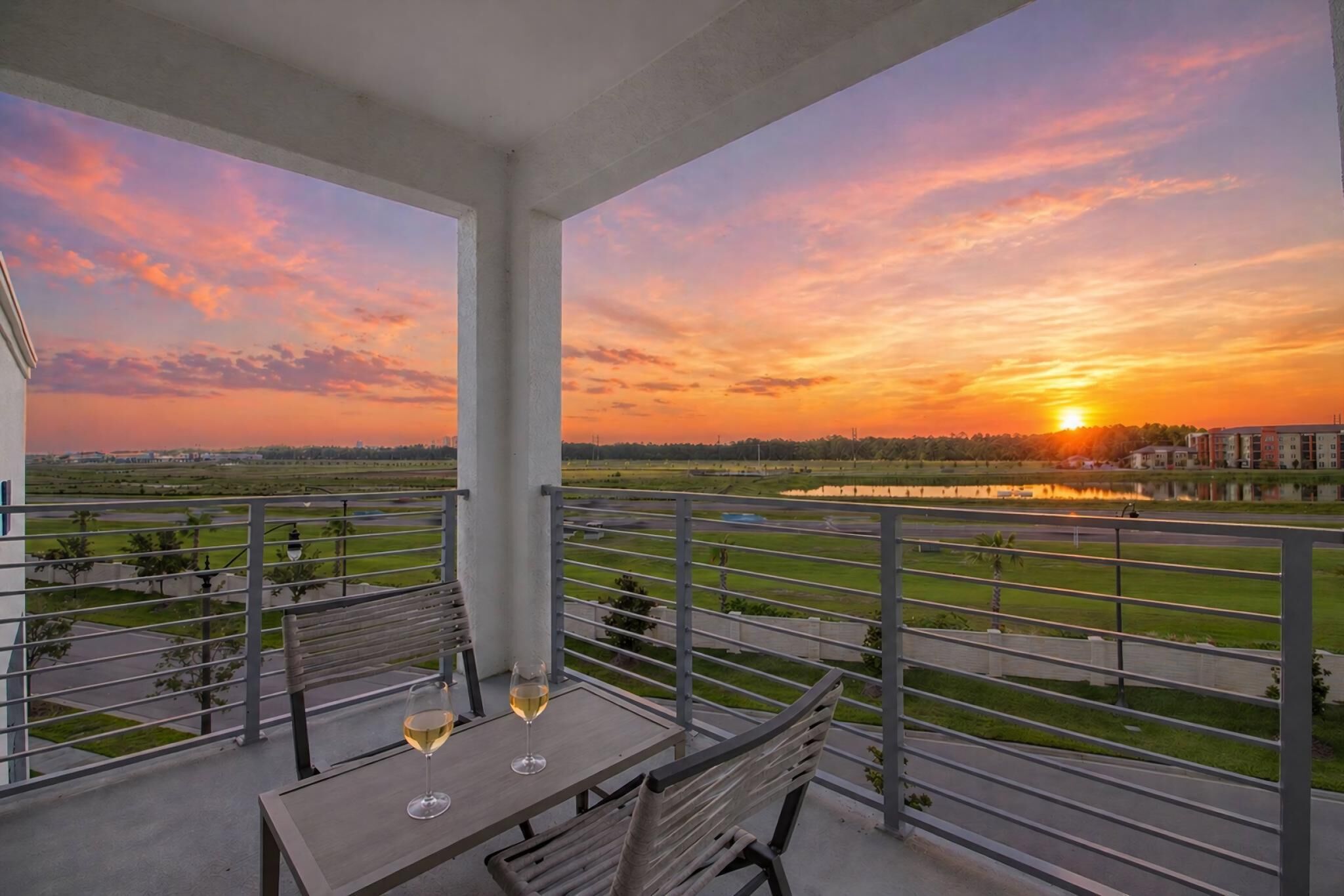 Ein Balkon mit Tisch und Stühlen, der Blick auf einen Sonnenuntergang über ein Gewässer und entfernte Gebäude bietet.