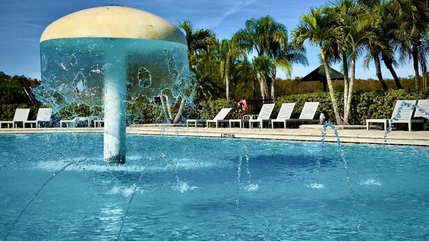 A swimming pool with a large water feature, surrounded by lounge chairs and palm trees. - 2633 Trafalgar Village (Kissimmee)