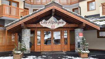 Een hotelgevel met een bordje waarop “Les Alpages” staat, houten balkons, stenen zuilen en een met sneeuw bedekte grond.