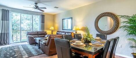 A spacious living room with a dining area, large windows, and a ceiling fan.