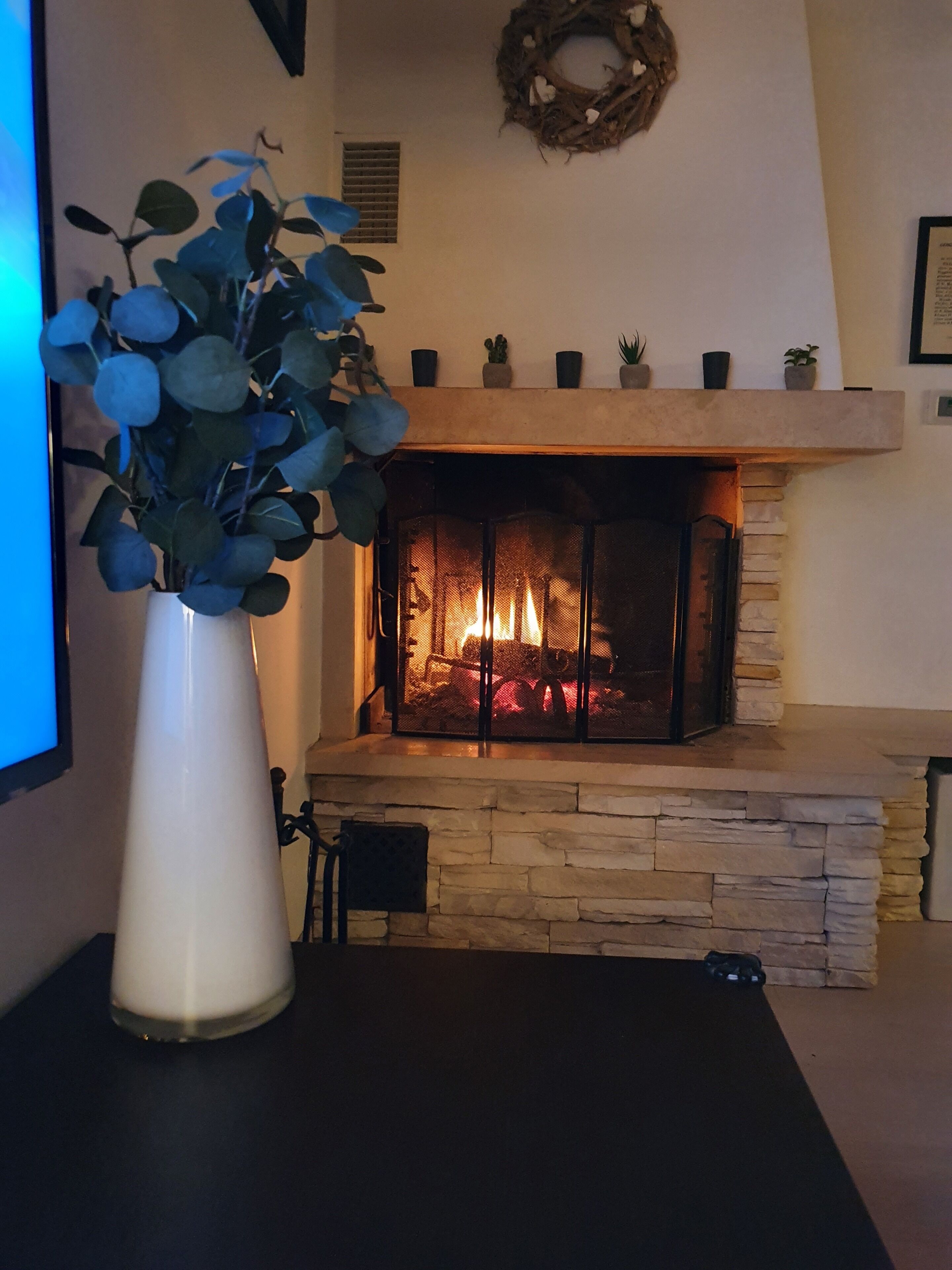 Una chimenea con revestimiento de piedra, un florero blanco con hojas azules y una pequeña mesa con una silla frente a ella.