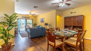 A spacious living room with a sofa, dining table, and chairs. There is a ceiling fan, a large plant, and a sign that reads "BEACH". - 8118 Windsor Palms (Kissimmee)