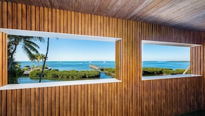 A room with wooden panel walls and two windows with white frames, each offering a view of a coastal landscape with palm trees and a pier. - Iconic Ted Williams Estate. Bayfront, Heated Infinity Pool & Hot Tub, Dockage. (Islamorada)