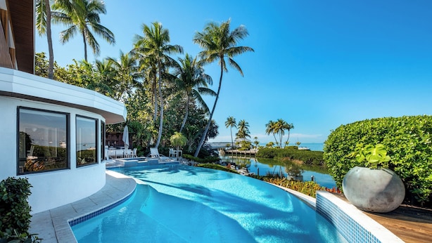 A modern pool area with clear blue water, surrounded by palm trees and a view of the ocean. - Iconic Ted Williams Estate. Bayfront, Heated Infinity Pool & Hot Tub, Dockage. (Islamorada)