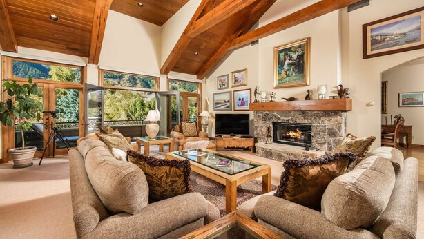 A spacious living room with a stone fireplace, wooden beams, and large windows offering a view of the outdoors. - Homes (Aspen)