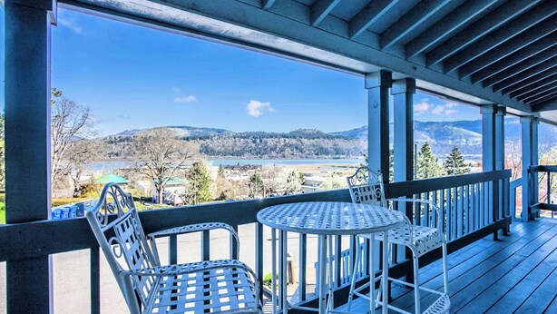 A covered outdoor seating area with metal chairs and a table, overlooking a scenic landscape with hills and a body of water. - Enjoy the View - 2 Bedrooms, 2 Bathrooms - Upstairs Condo Blocks from Downtown (Hood River)