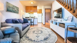 A modern living room with a sofa, a coffee table, and a patterned rug. There is a kitchen area in the background with a dining table and chairs. A staircase with wooden balusters is visible on the right side. - Enjoy the View - 2 Bedrooms, 2 Bathrooms - Upstairs Condo Blocks from Downtown (Hood River)