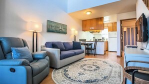 A modern living room with a sofa, a coffee table, and a rug. There is a kitchen area in the background with wooden cabinets and a dining table. - Enjoy the View - 2 Bedrooms, 2 Bathrooms - Upstairs Condo Blocks from Downtown (Hood River)