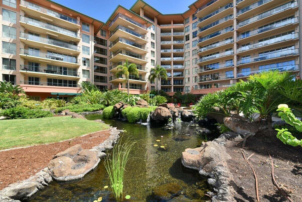 A hotel with multiple balconies, a landscaped garden with a small pond, and a variety of tropical plants.
