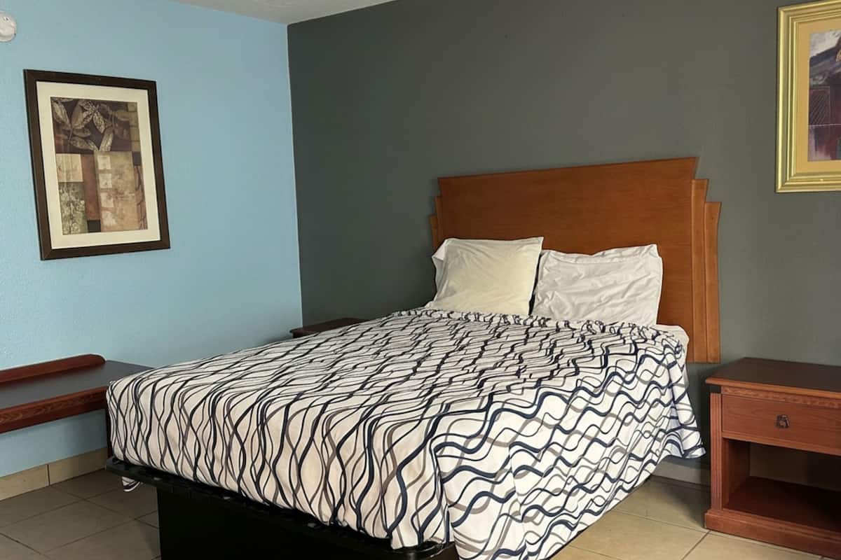 A bedroom with a bed, a nightstand, a wooden headboard, a framed picture on the wall, and a window.