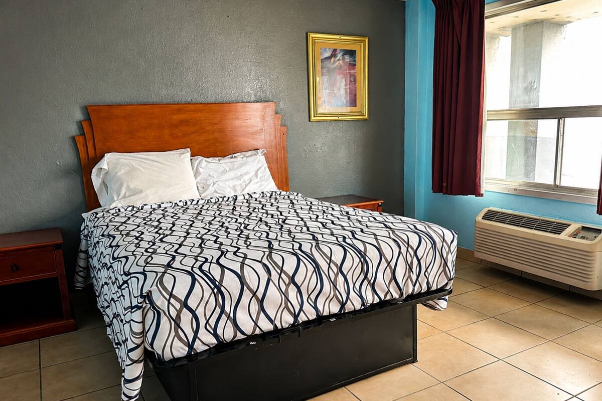 A bedroom with a bed, nightstands, a window with curtains, a wall-mounted air conditioner, and a framed picture.