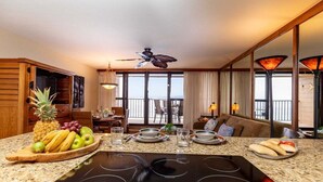 A modern kitchen with a large island, a dining area with a view, and a living area with a sofa and a TV.