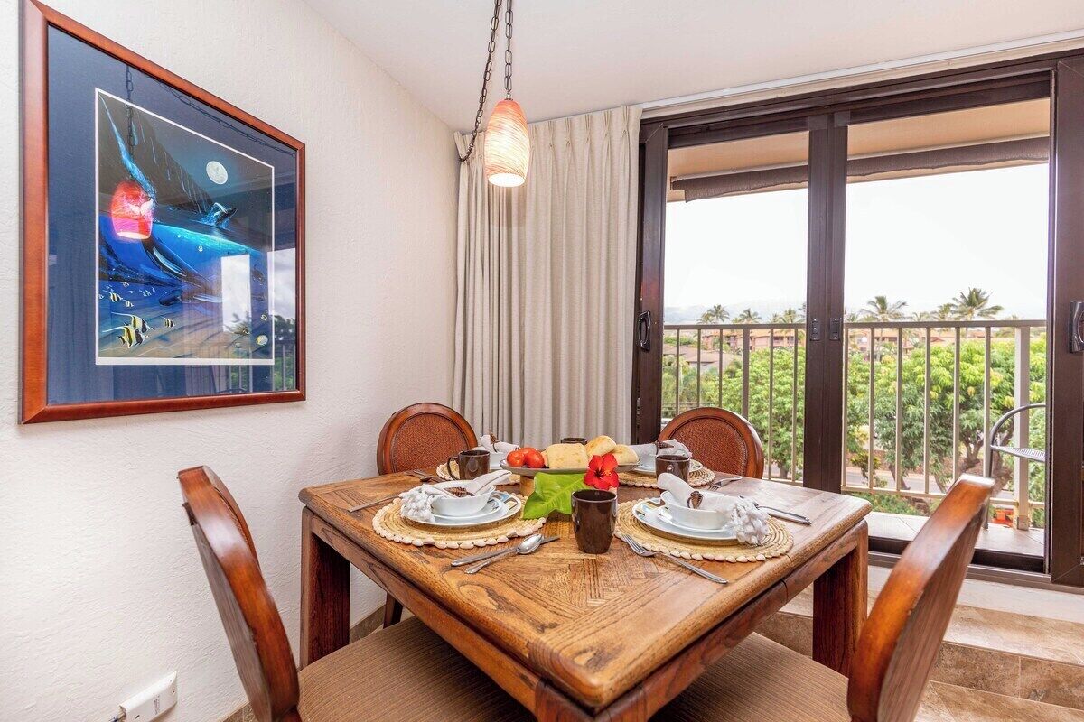 A dining area with a wooden table set for four, a framed picture on the wall, and a balcony with a view of greenery.