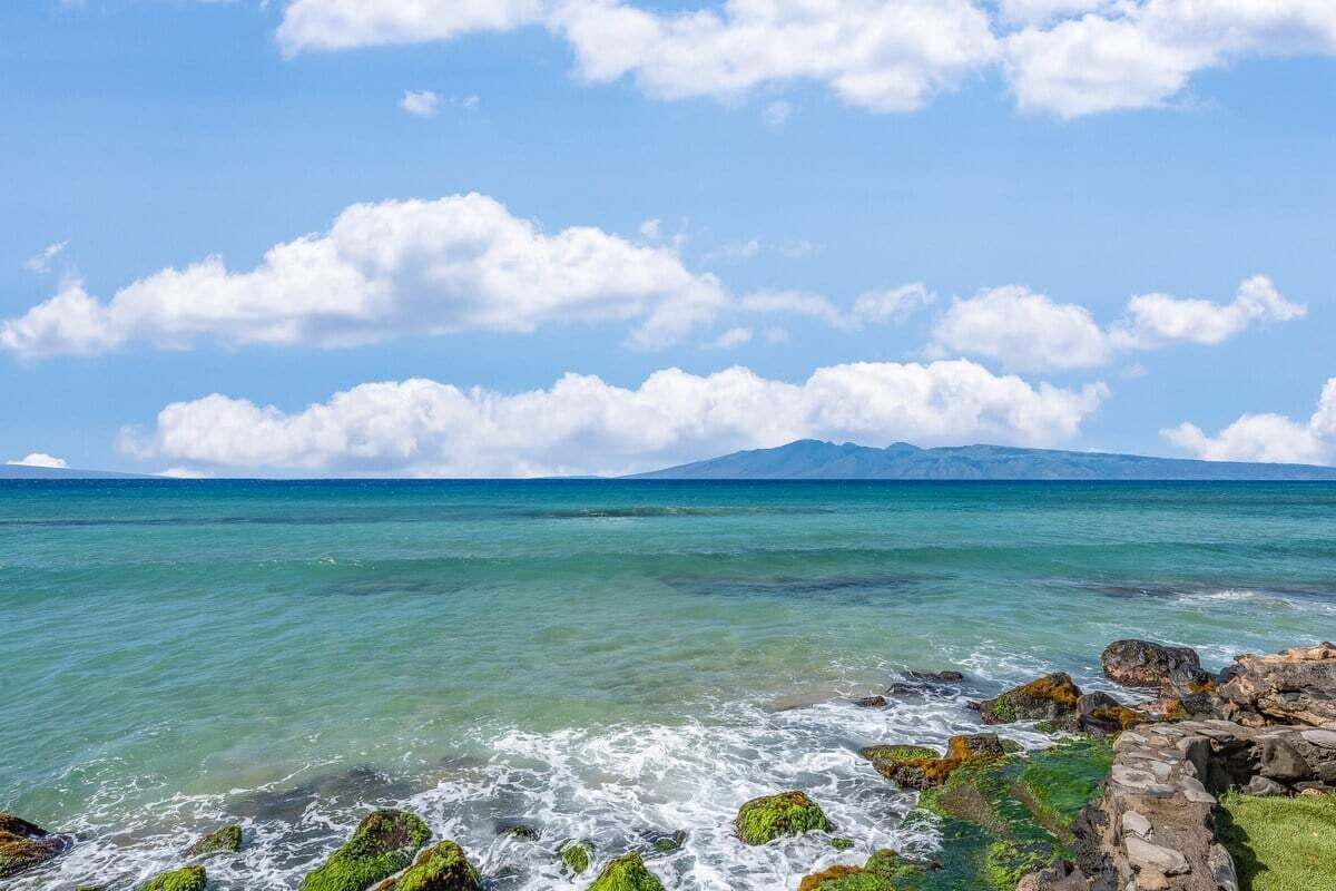 A coastal landscape with clear turquoise waters, rocky shore, and distant mountains under a partly cloudy sky.
