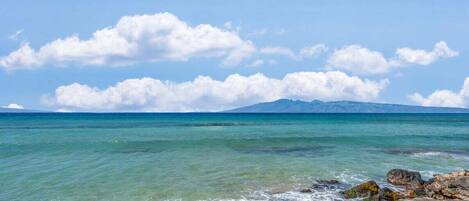 A coastal landscape with clear turquoise waters, rocky shore, and distant mountains under a partly cloudy sky.