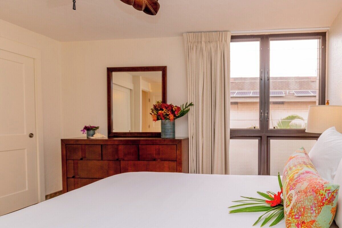 Une chambre d’hôtel avec un lit, une commode en bois, un miroir, une fenêtre avec des rideaux et une vue sur les bâtiments environnants.