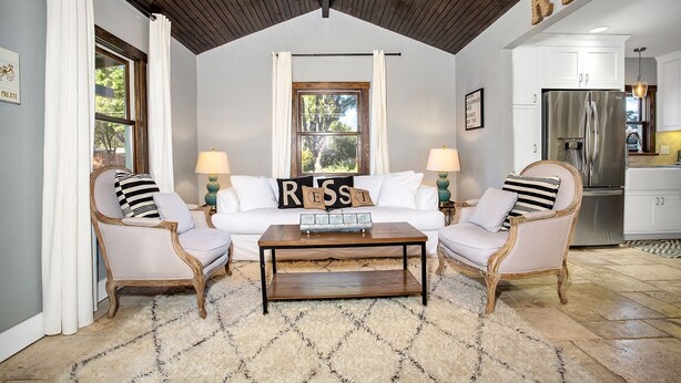 A living room with a wooden ceiling, a white sofa with 'RESS' pillows, two armchairs, a coffee table, and a kitchen in the background. - Rest Haus | In Town | Hot Tub (Fredericksburg)