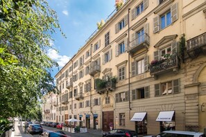 Una hilera de edificios de varios pisos con balcones, locales comerciales y coches estacionados en un día soleado.