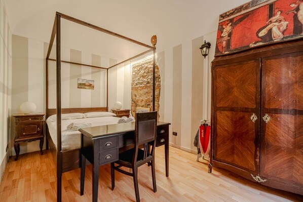 Una cama con dosel y cabecera, un escritorio con cajones, una silla, un armario con espejo y una lámpara de pared.