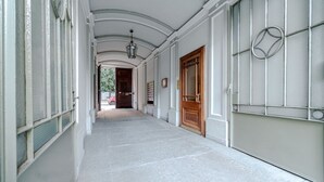 Un couloir avec une porte en bois, une fenêtre ornée d’une grille métallique et un luminaire suspendu.