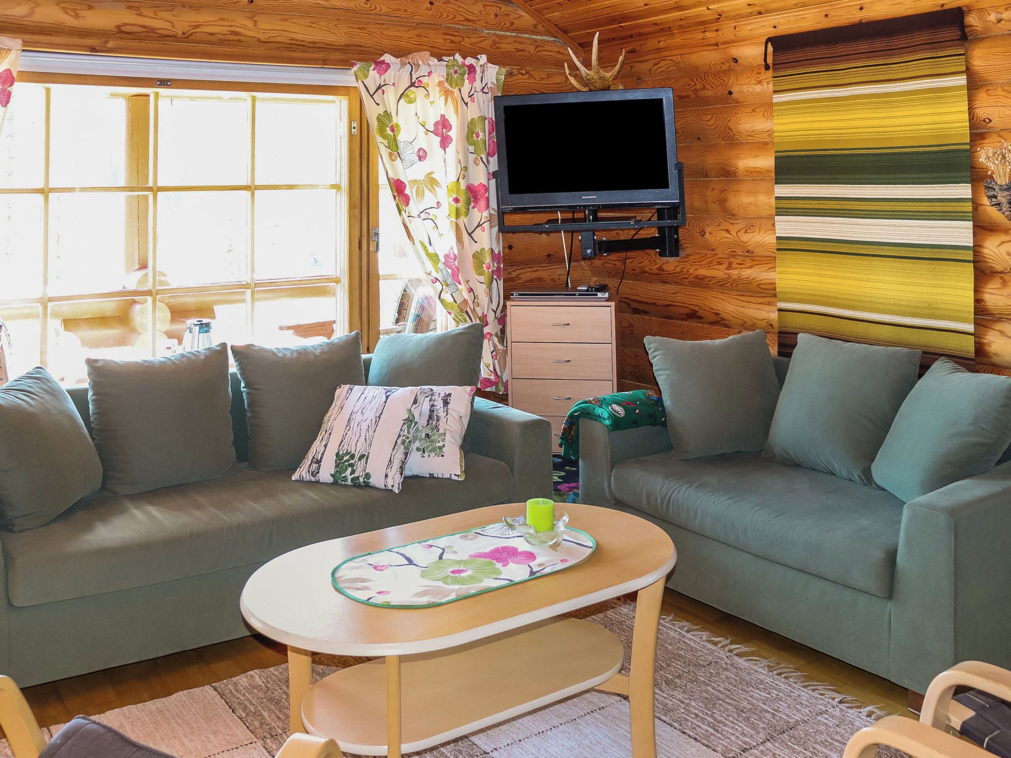 L’intérieur chaleureux d’un chalet avec deux canapés, une petite table, un téléviseur fixé au mur et une fenêtre avec des rideaux.