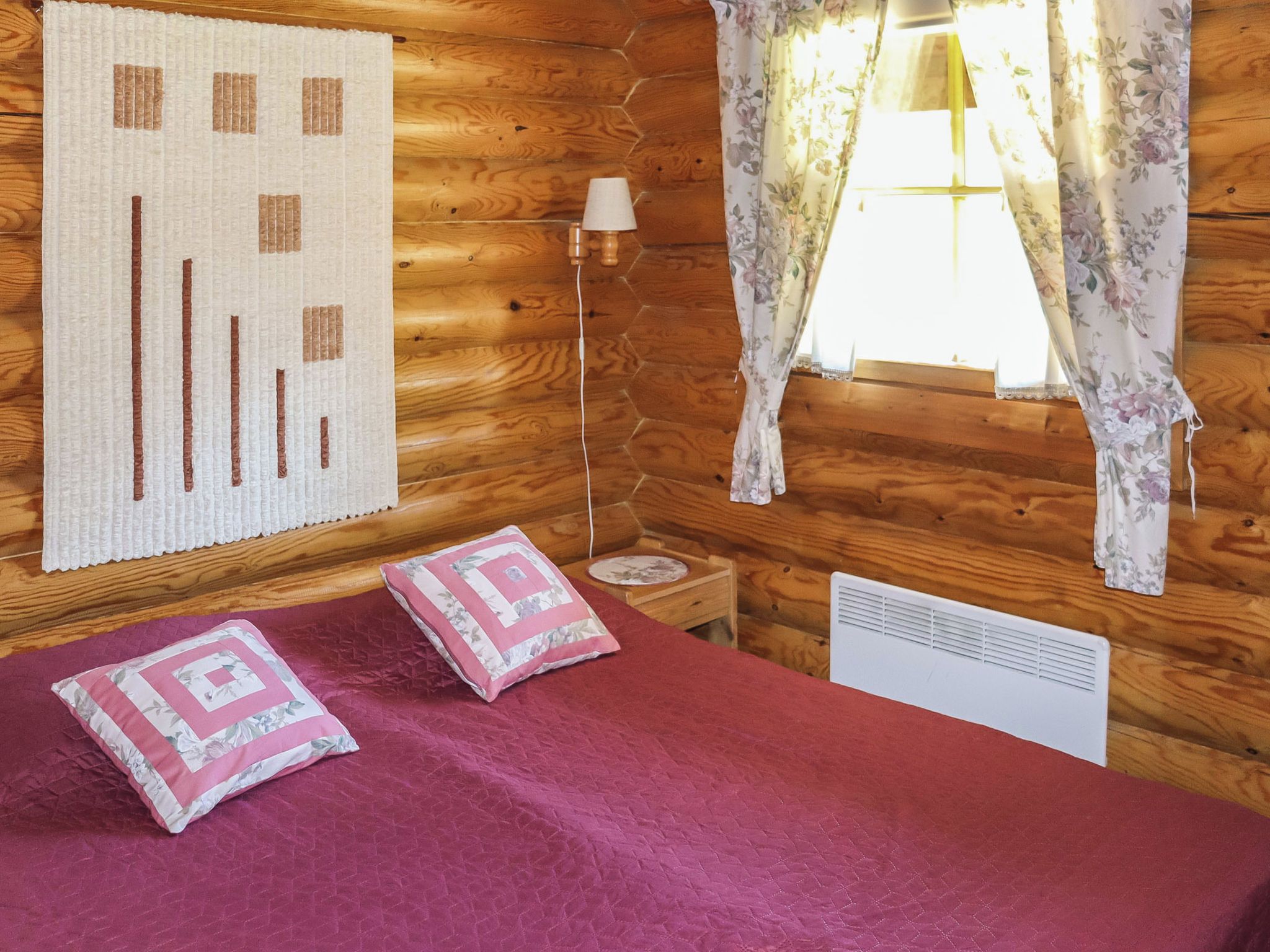A wooden cabin room with a bed, a lamp, a window with floral curtains, and a wall hanging.