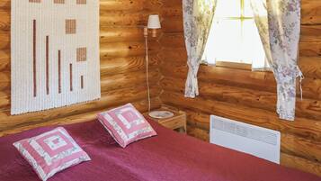 A wooden cabin room with a bed, a lamp, a window with floral curtains, and a wall hanging.