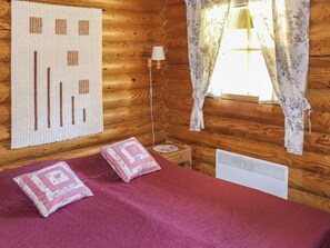A wooden cabin room with a bed, a lamp, a window with floral curtains, and a wall hanging.