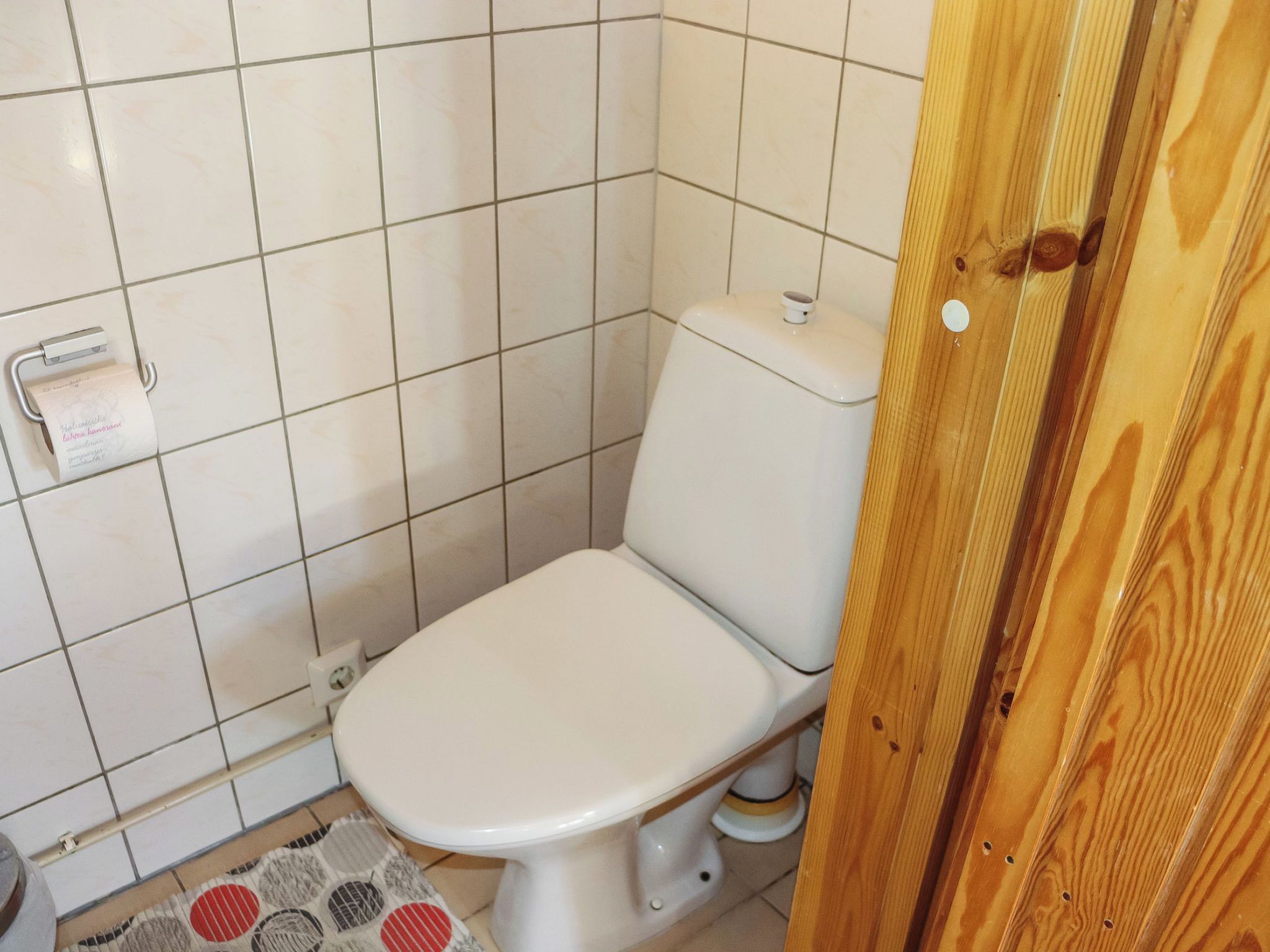A small, tiled bathroom with a toilet, a paper towel dispenser, and a wooden door.