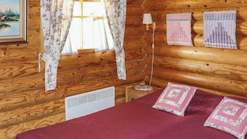 A wooden cabin room with a bed, two pillows, a lamp, and a window with floral curtains.