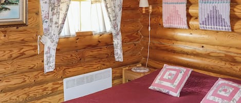 Ein Zimmer einer Holzhütte mit einem Bett, zwei Kissen, einer Lampe und einem Fenster mit floralen Vorhängen.