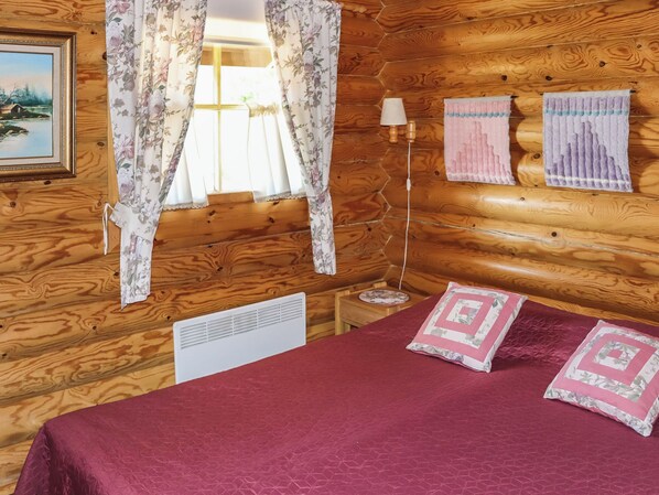 A wooden cabin room with a bed, two pillows, a lamp, and a window with floral curtains.