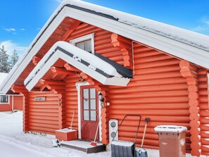A red log cabin with a snow-covered roof, a front door, and a small porch area with a broom and shovel.
