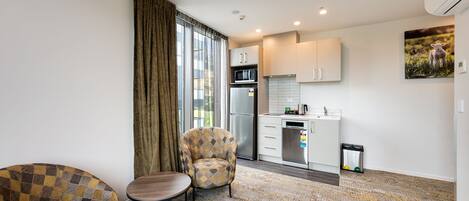 A modern living room with a sofa, armchair, and a small table. A kitchen area is visible with a refrigerator, microwave, and sink.