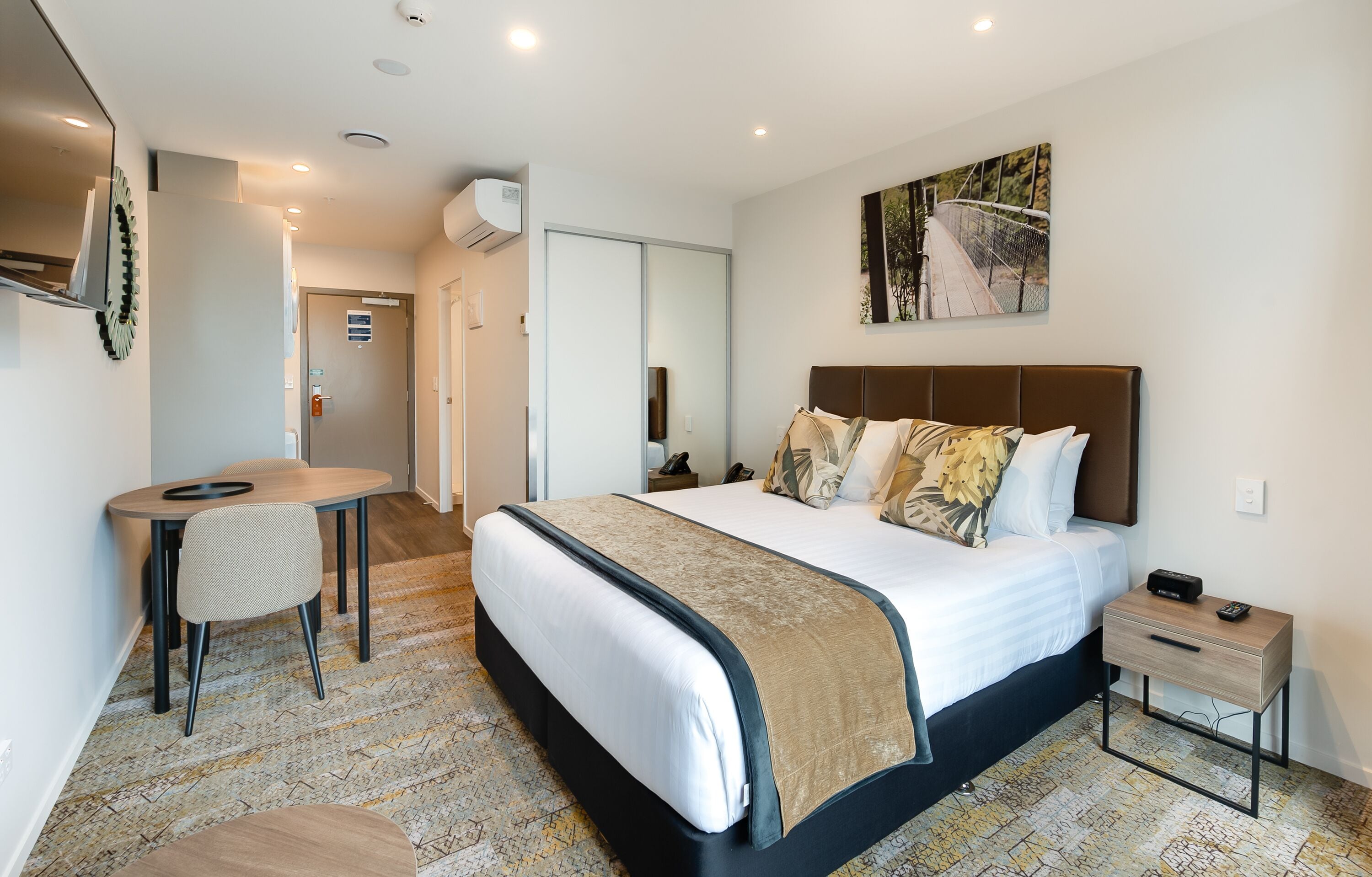 A modern hotel room with a large bed, a desk, and a chair. There is a painting on the wall and a small table with a remote control.