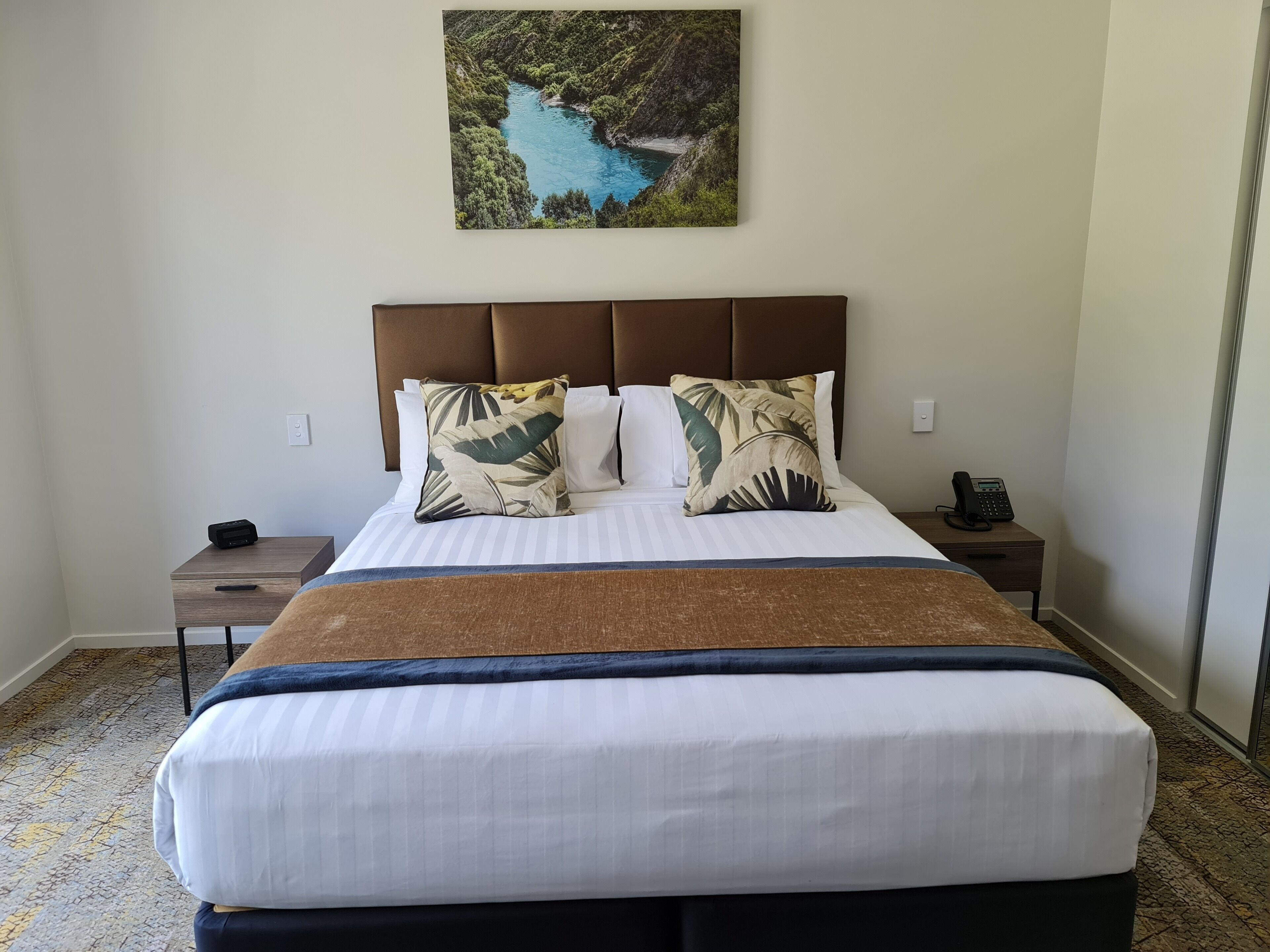 Una cama bien hecha con una colcha a rayas blanca y azul, dos almohadas con motivos de hojas tropicales y un cabecero marrón. Se ve una mesita de noche con un teléfono y un control remoto.