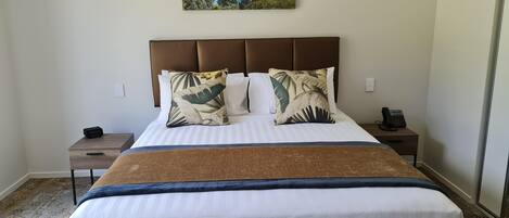 A neatly made bed with a striped white and blue bedspread, two pillows with tropical leaf patterns, and a brown headboard. A small bedside table with a phone and a remote control is visible.