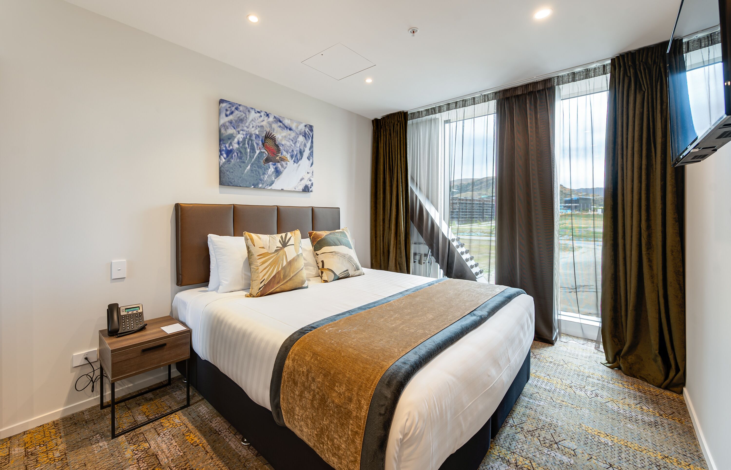 Een hotelkamer met een groot bed, een nachtkastje met een telefoon, een schilderij aan de muur en uitzicht op het omliggende landschap door het raam.