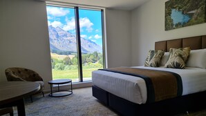 Une chambre d’hôtel avec un grand lit, une chaise, une petite table et une vue sur les montagnes par une grande fenêtre.