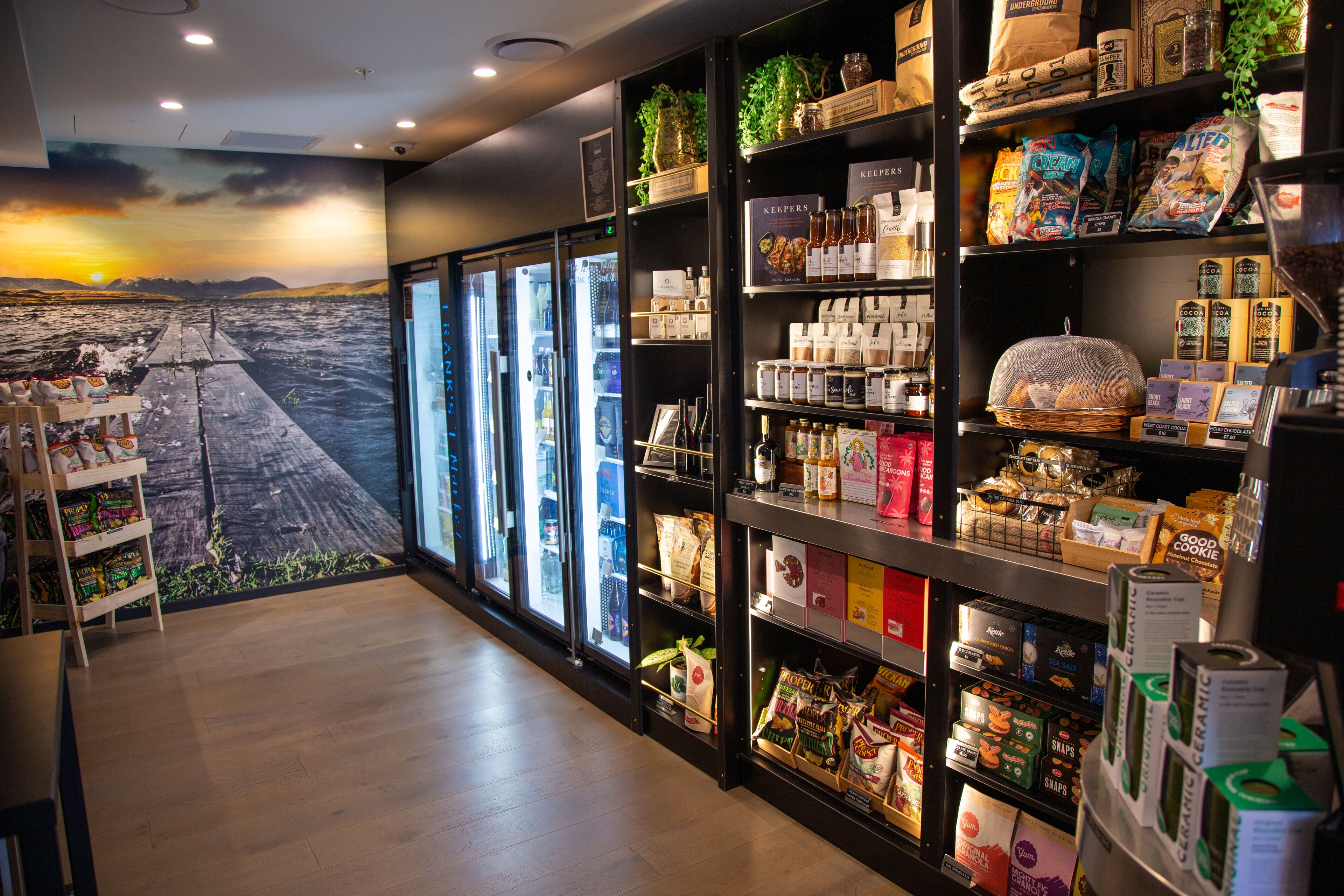 A store with shelves stocked with various products, a large window with a view of a sunset, and a wooden floor.