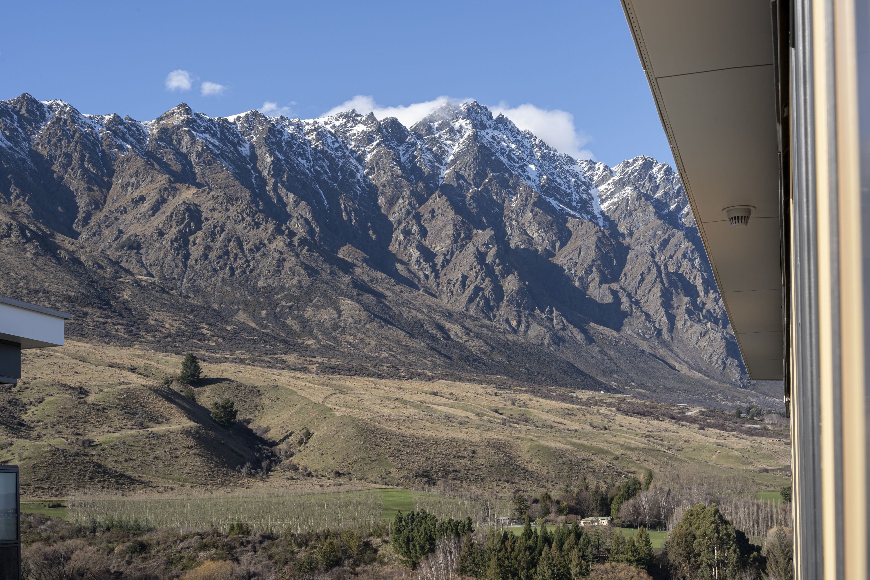 Paisagem montanhosa com picos nevados, um vale com vegetação rala e um edifício com telhado inclinado.