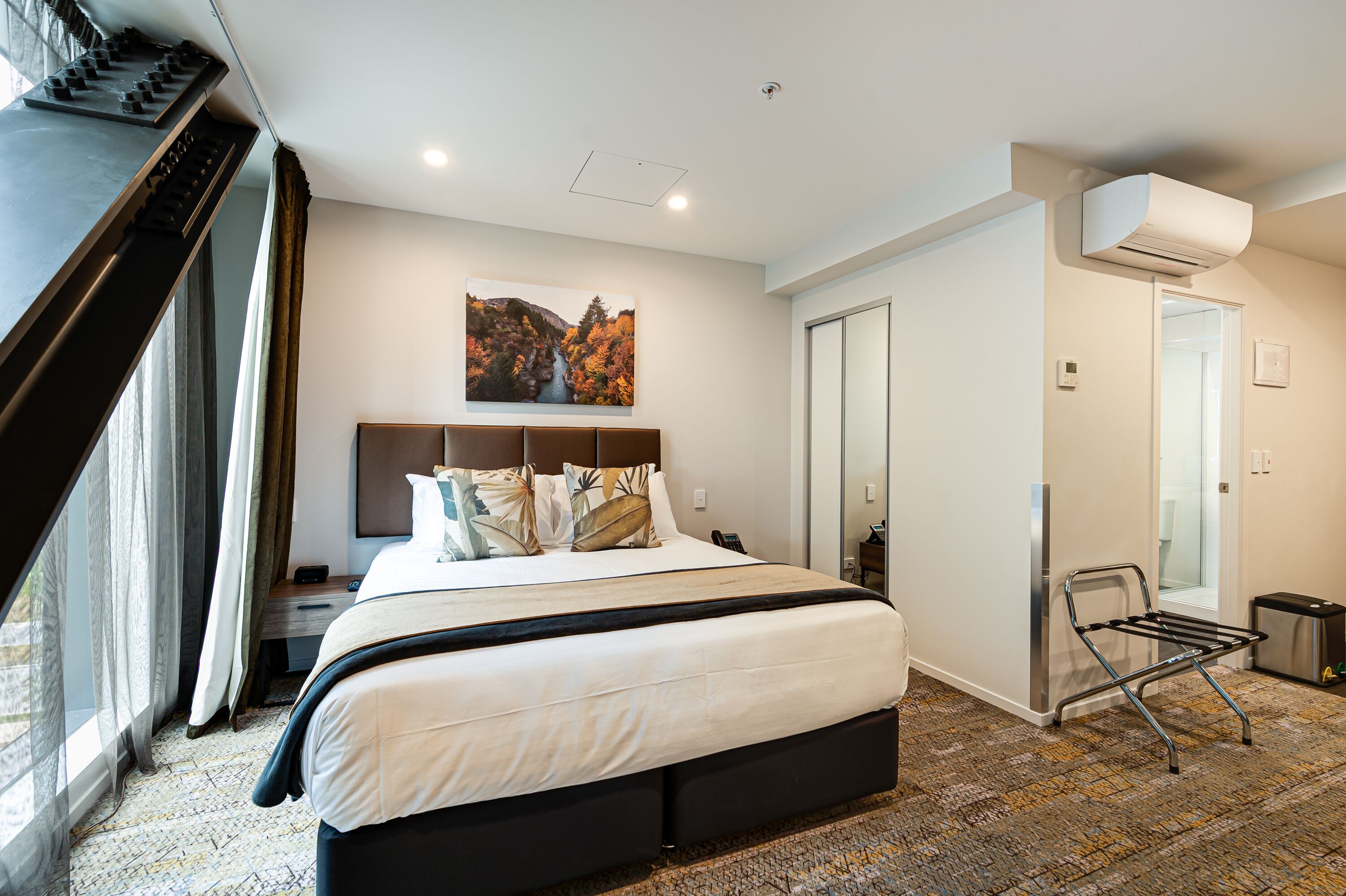 Habitación de hotel con una cama grande, un escritorio, una silla, una ventana con cortinas, un equipo de aire acondicionado y un cuadro en la pared.