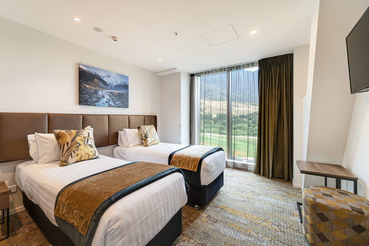 Ein Hotelzimmer mit zwei Betten, einem großen Fenster mit Vorhängen, einem Flachbildfernseher und einem Gemälde an der Wand.