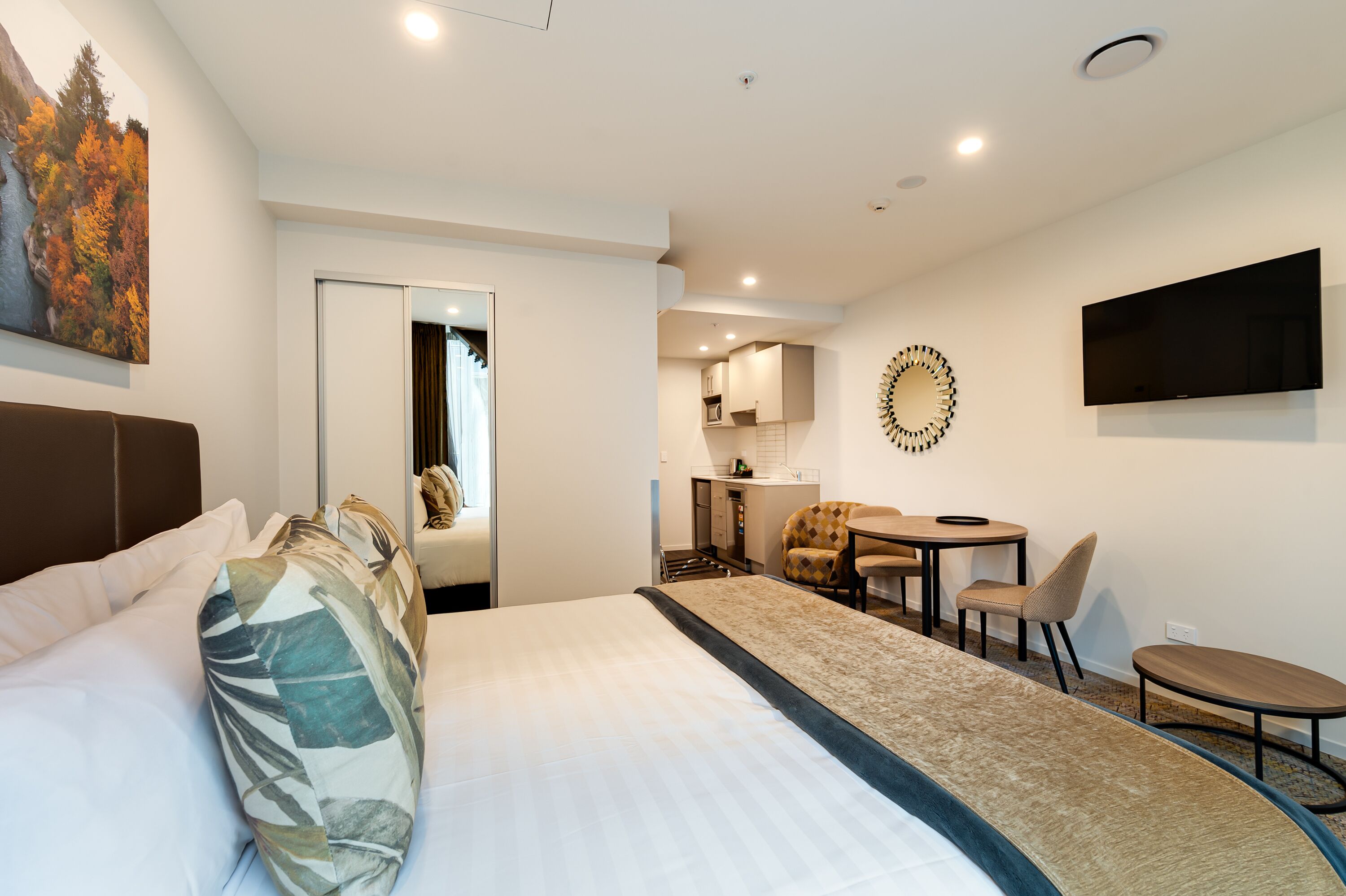 A hotel room with a bed, a desk, a chair, a TV, and a kitchen area.