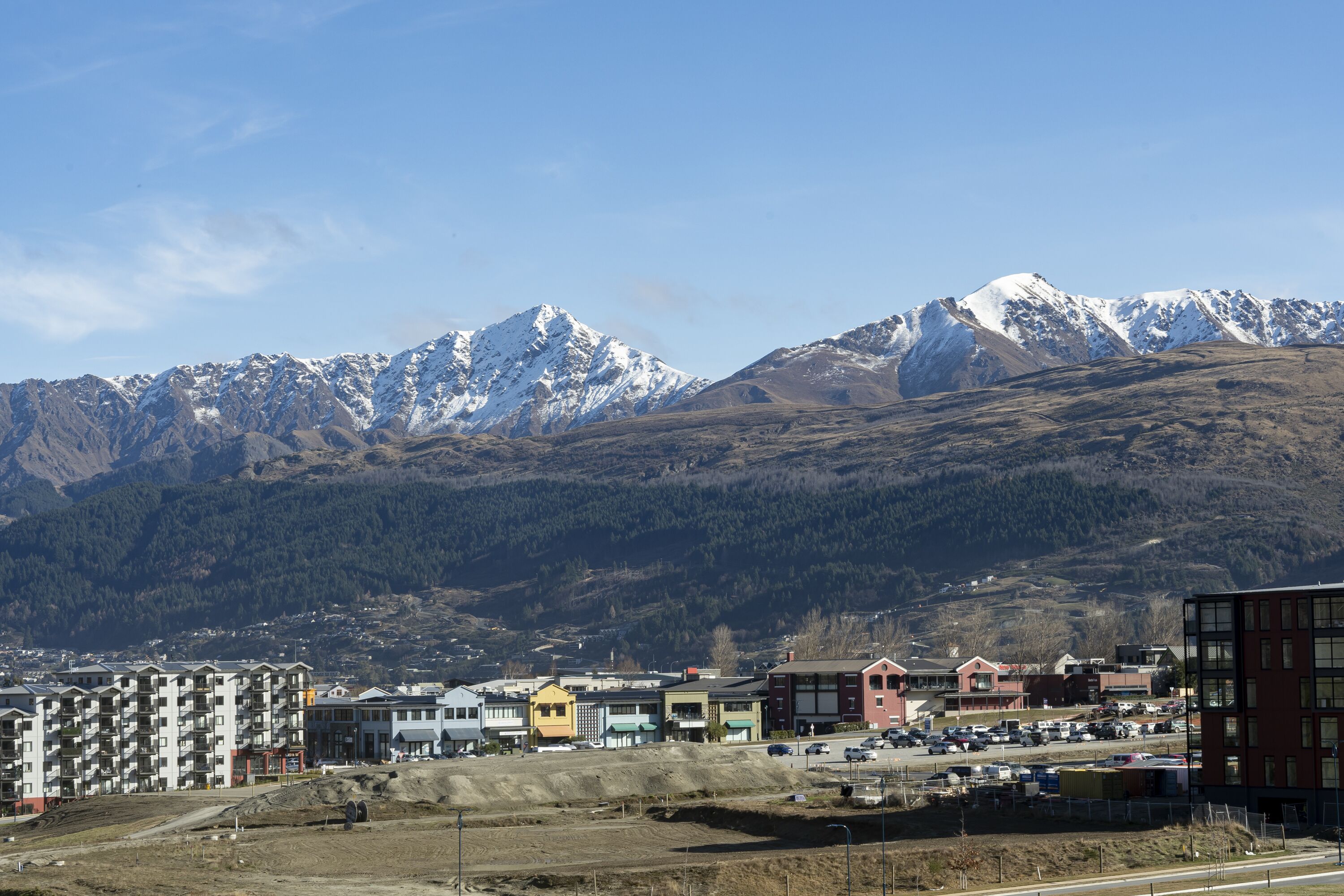 A town with a mountainous backdrop, featuring multi-story buildings, a parking lot, and a construction site.